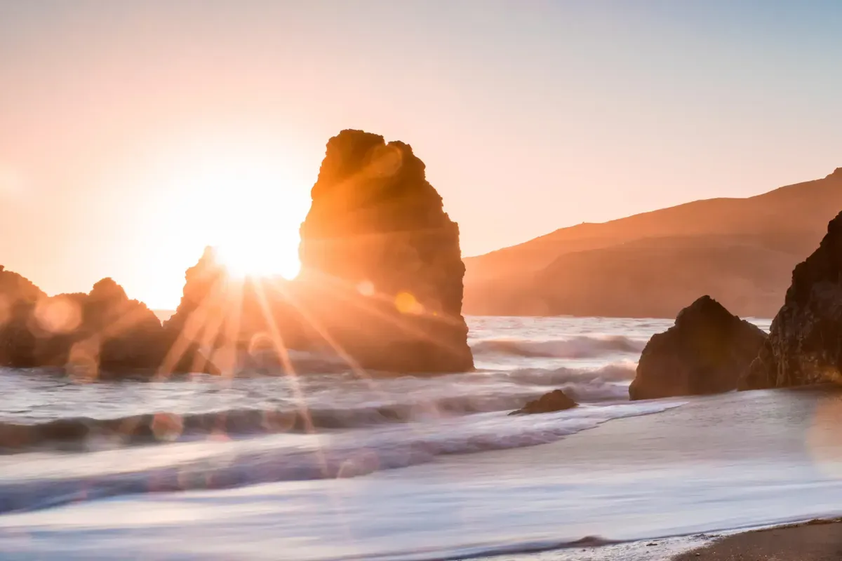 Sun rays breaking through rocky sea stacks at a beach sunset