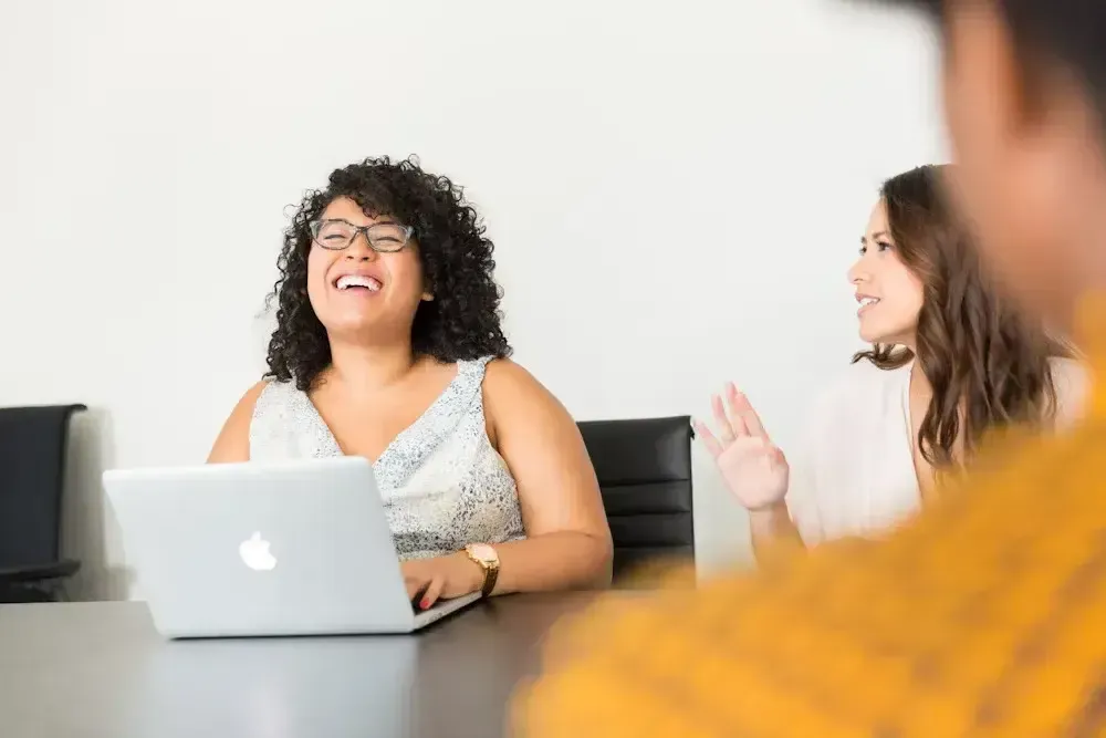 woman laughing at her laptop while a colleague speaks nearby