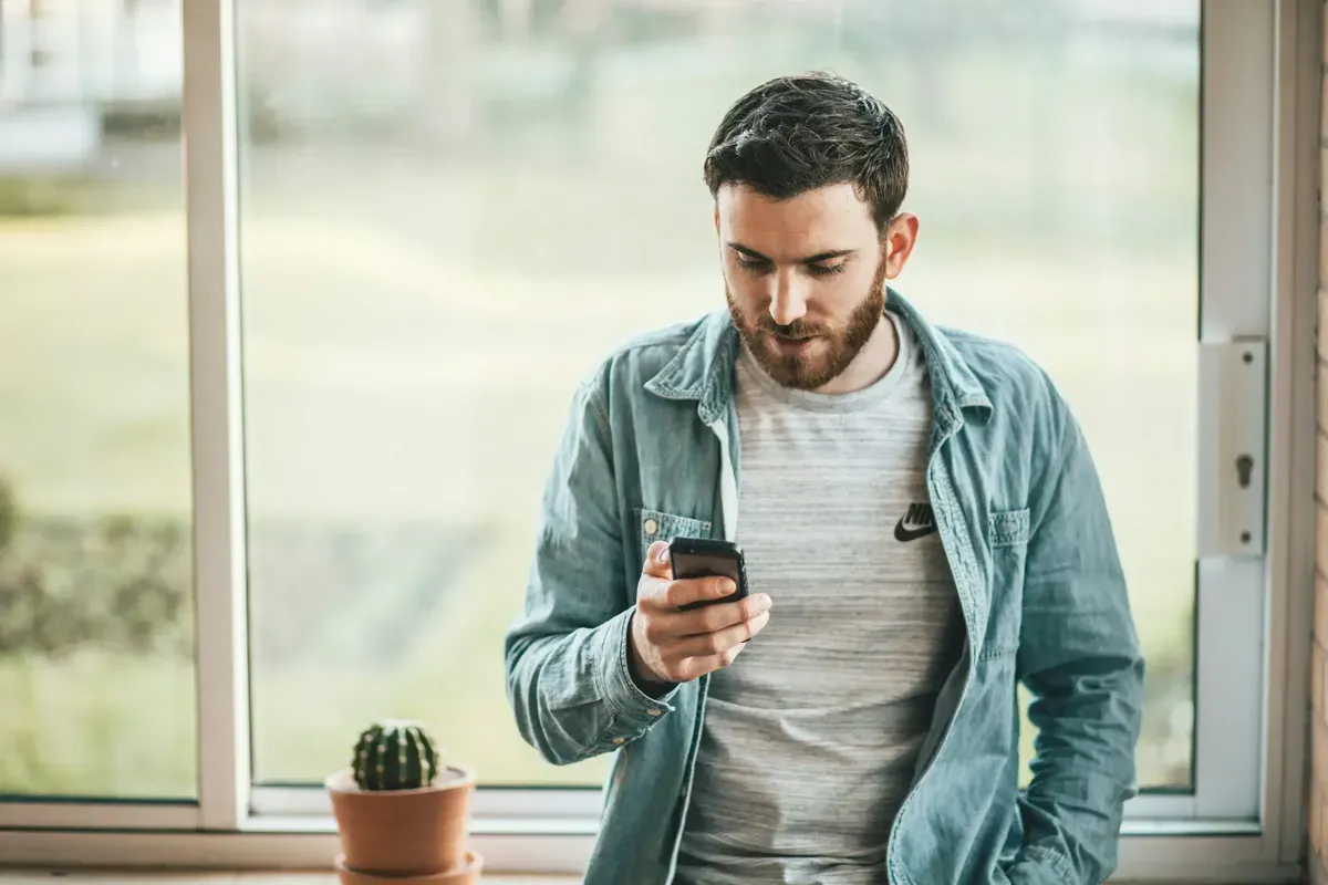 Man checking his phone near a window with a small cactus