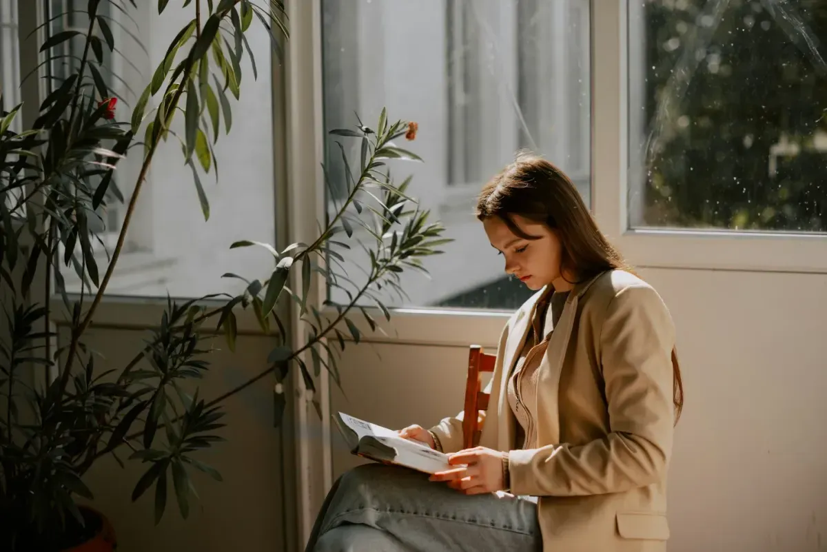 Woman reading a book by a sunlit window surrounded by houseplants