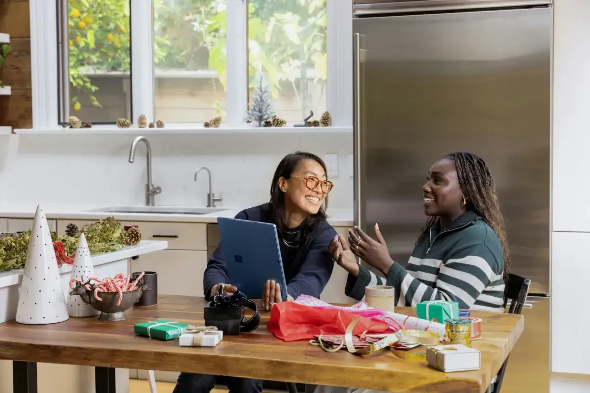 two women laughing while wrapping gifts with a laptop nearby